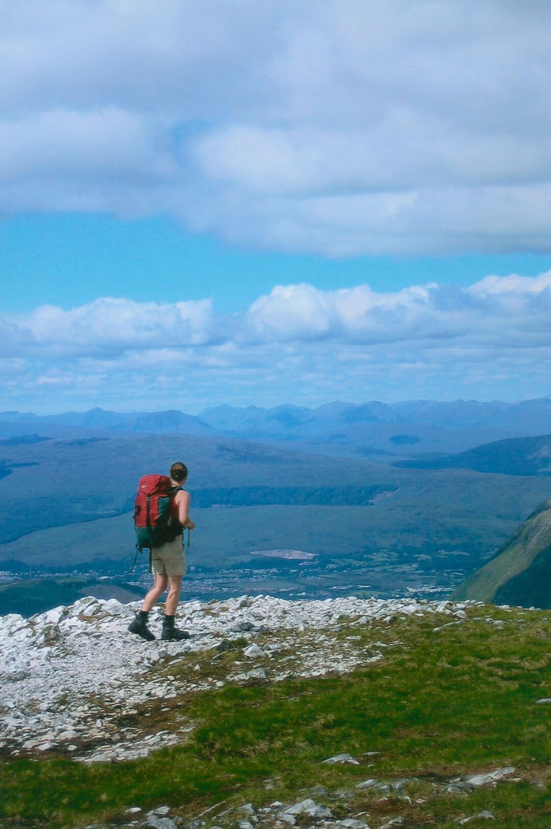 Sheryl Bartlett carries a red backpack and stands on top of a mountain facing out across the vista.