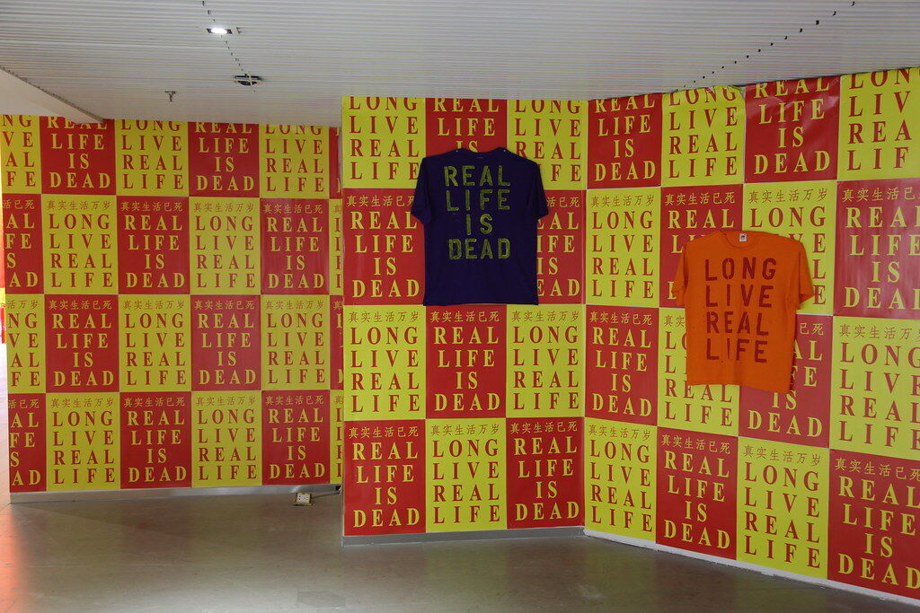 Bright yellow and orange posters cover the walls with a couple of t-shirts hung on the wall, the words 'long live real life' and 'real life is dead' are printed over both the posters and t-shirts.