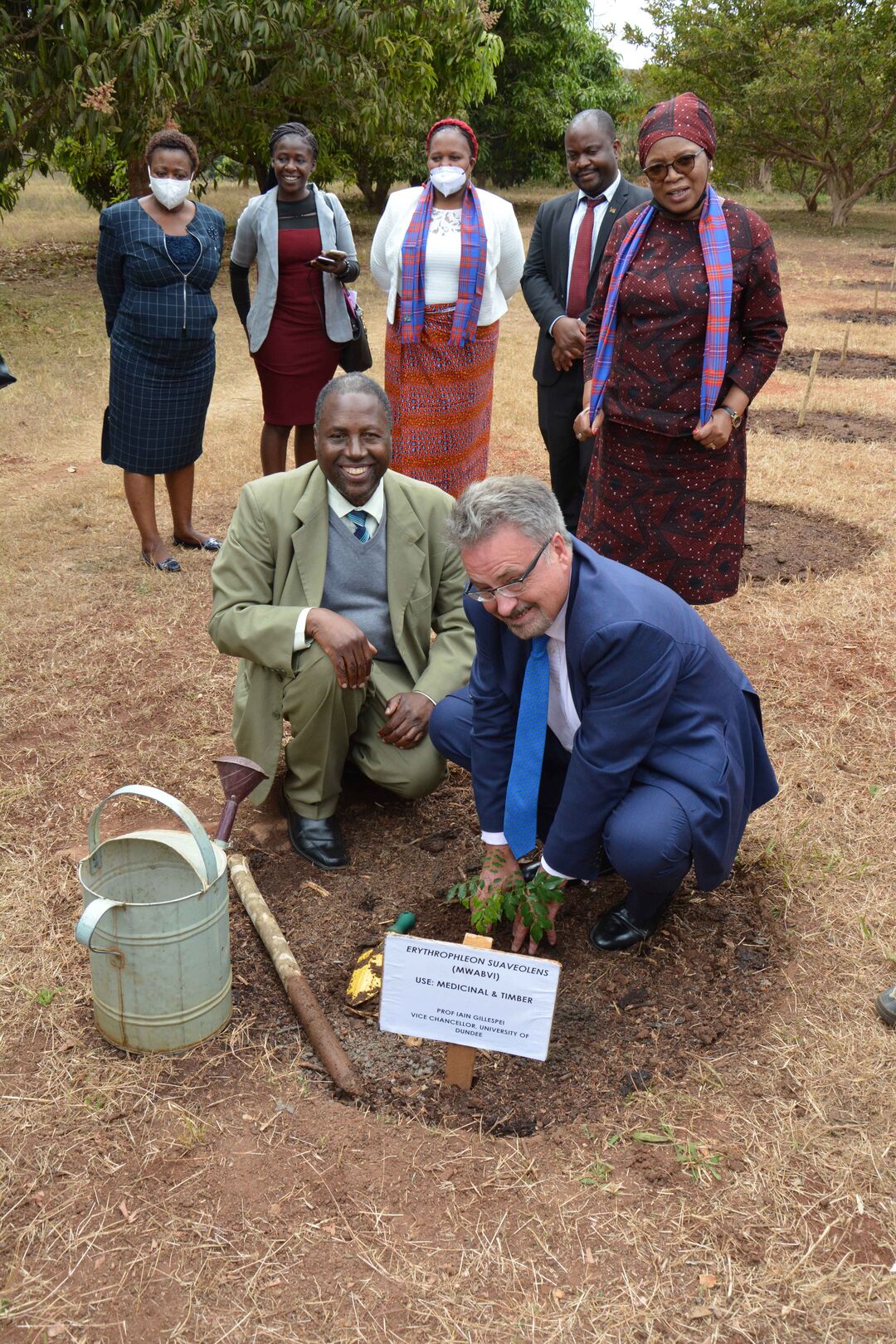 Principal Gillespie and Professor Chiotha crouch over a newly planted tree with a watering can to the left.