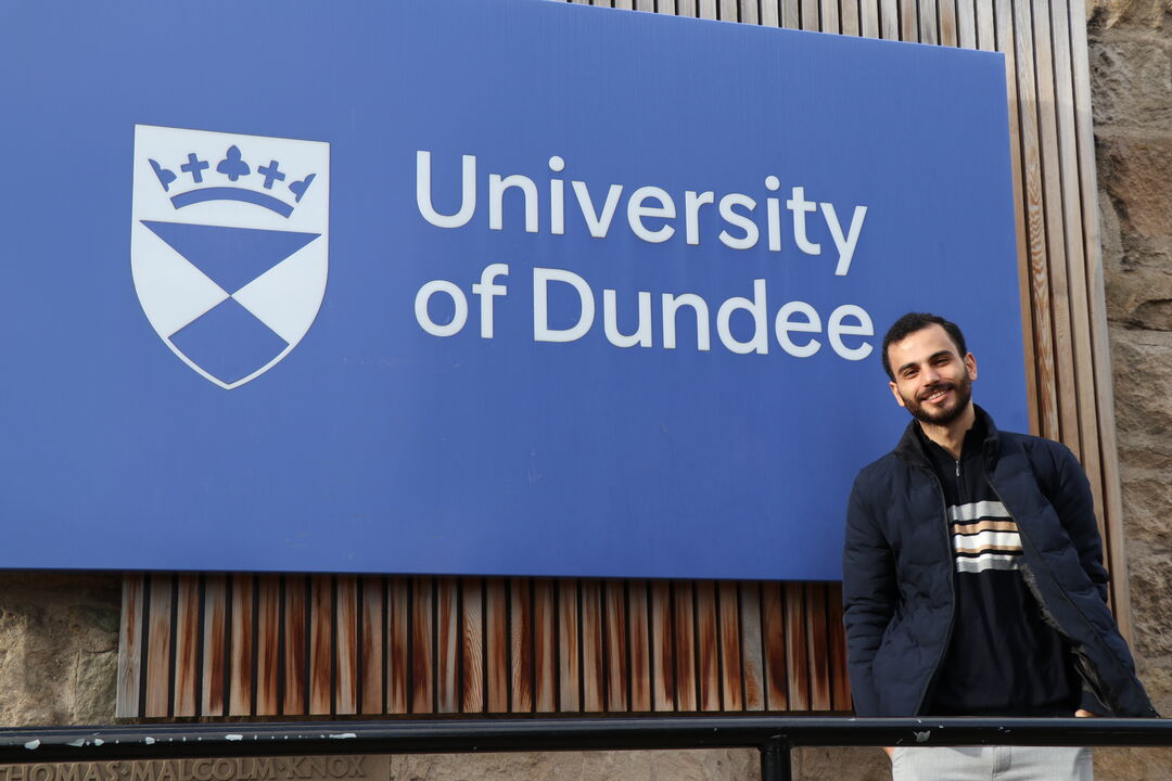 Bayan Alhasani standing in front of a large blue sign with 'University of Dundee' printed on it along with the University shield 