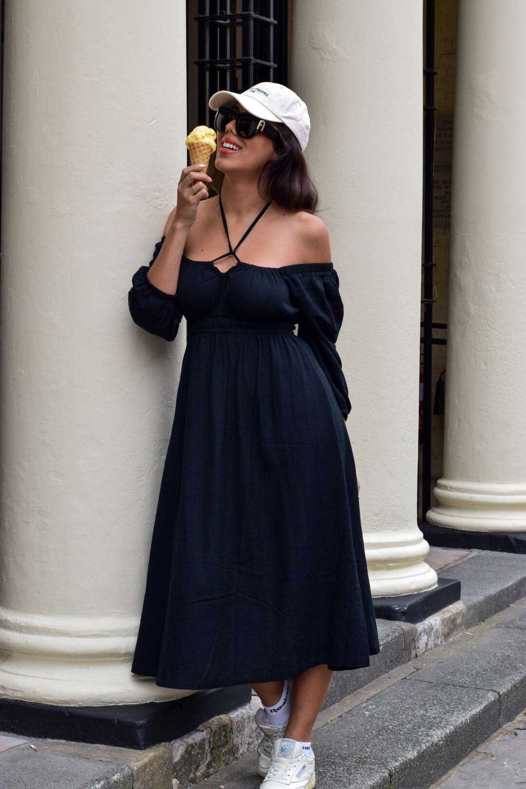 A model wearing a black dress with white trainers and a cap stands beside white pillars.
