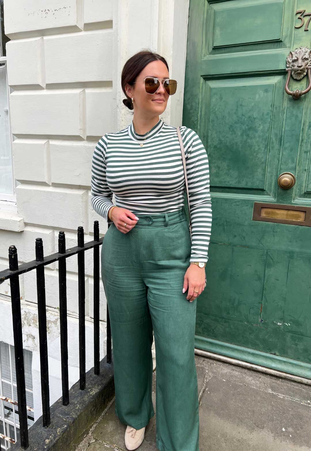 A model in green long sleeved top and trousers, standing on the step in front of a green door.