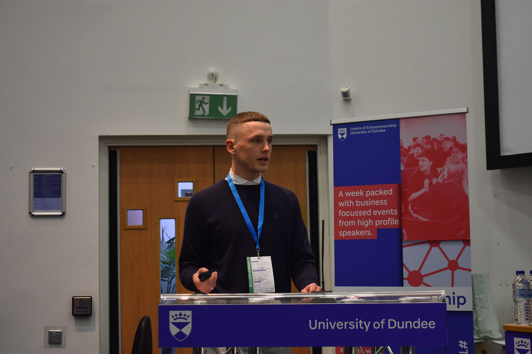 Robbie Beautyman presenting at the Venture 2019 competition standing at a podium with a blue Centre for Entrepreneurship banner