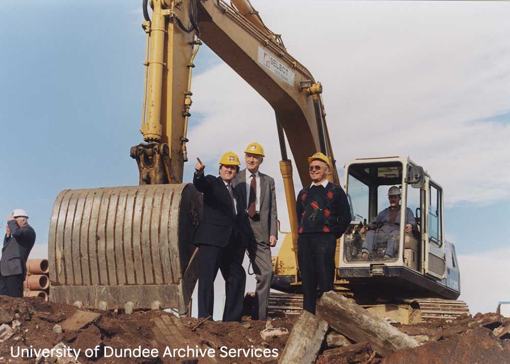 A man operating a digger, while more men stand on the dirt ground beside it.