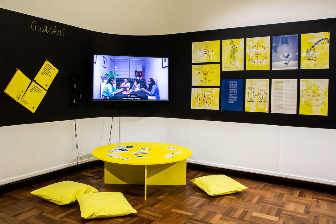 Exhibition installation view. TV, posters, yellow table and cushions by artistic collective GUDKSUL