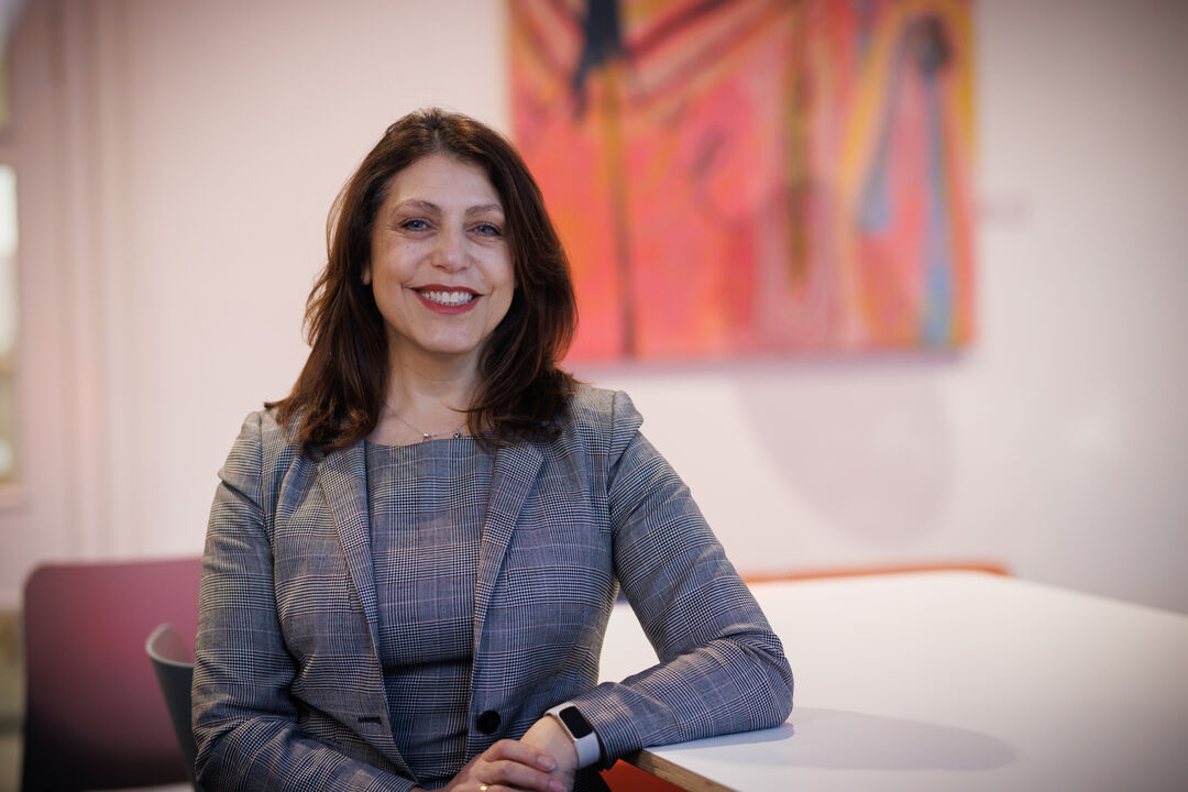 Woman wearing a grey suit sat on a chair with her elbow leaning on table to her side