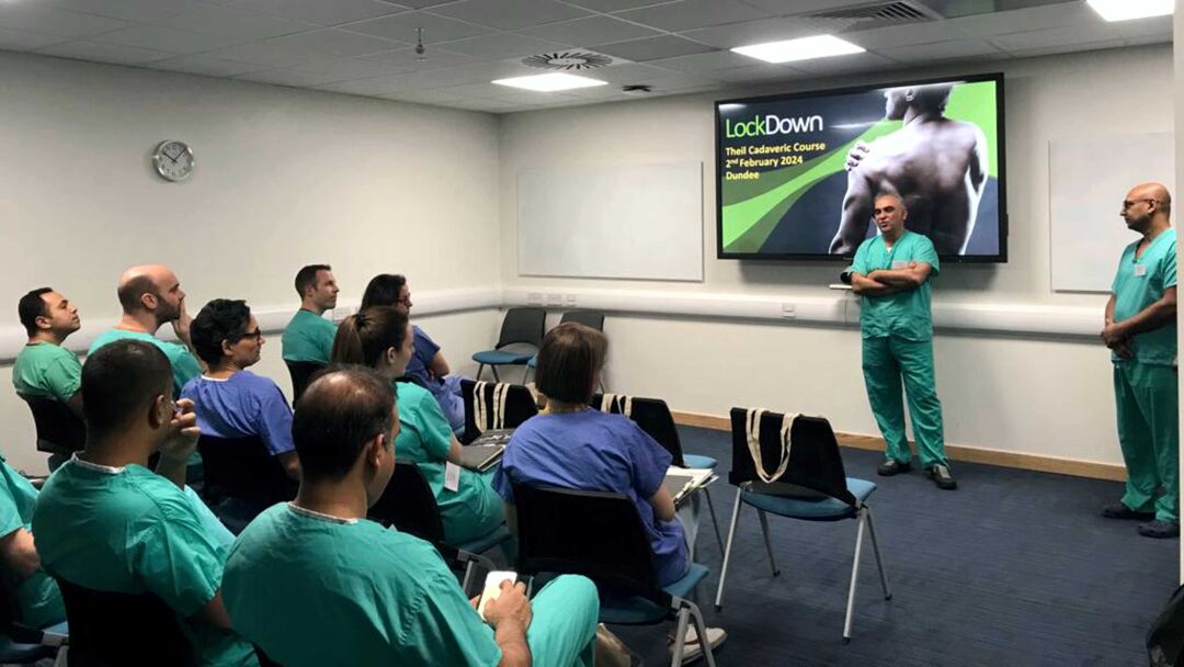 A man in surgeons uniform is presenting to a room full of other surgeons.