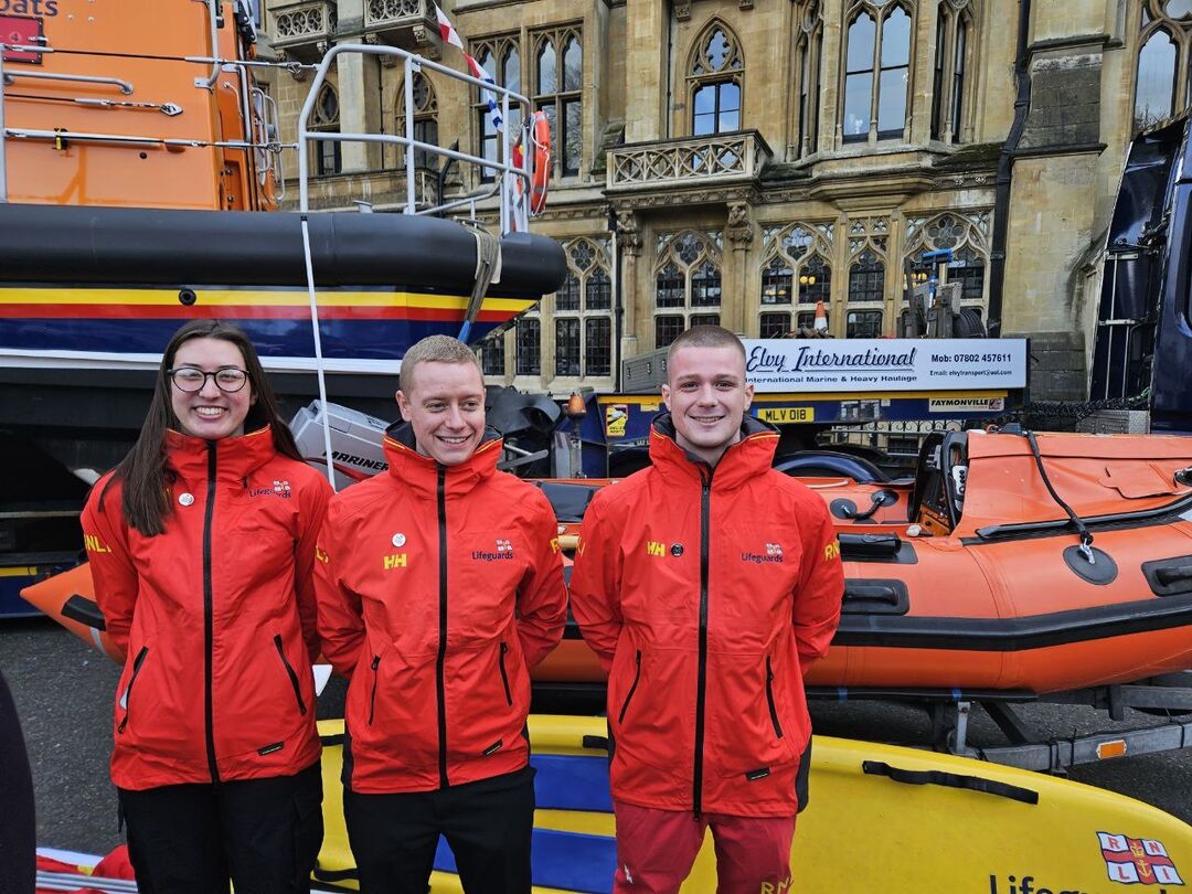 Three youths wearing red lifeguard jackets standing amongst boats parked on a street.