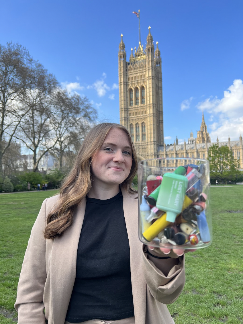 Woman holding a jar filled with discarded disposable vapes, standing outside Westminster
