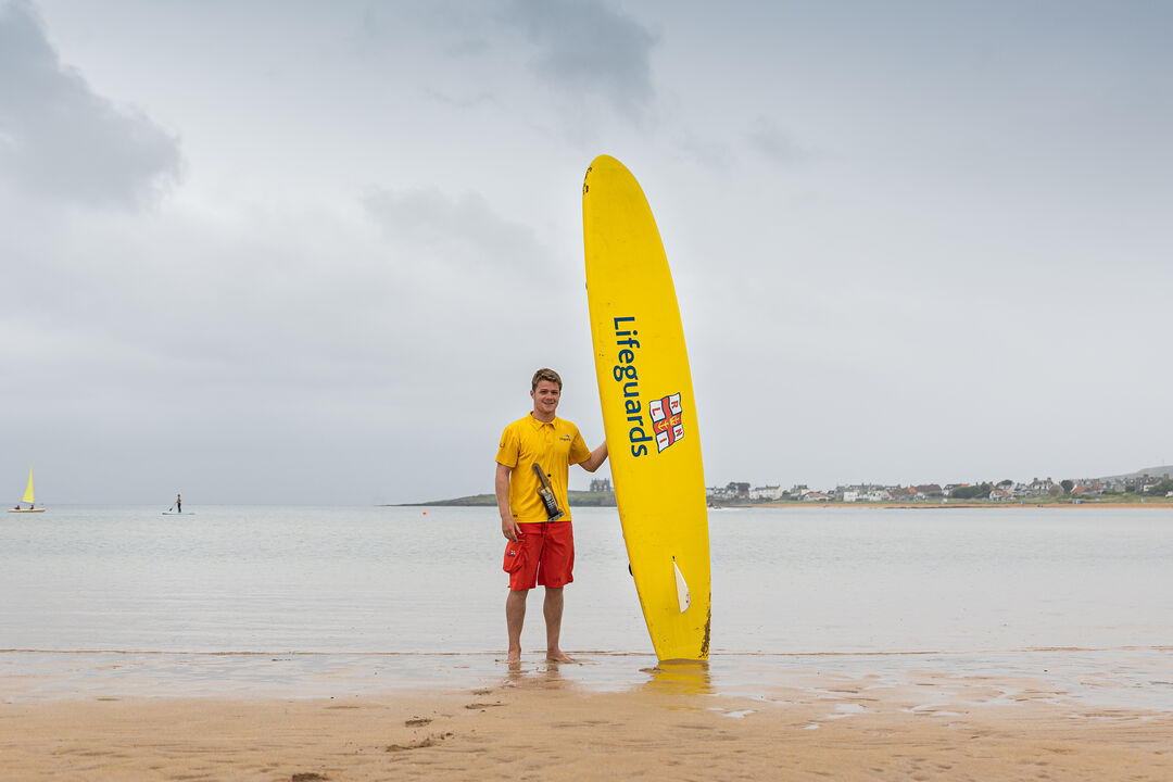 Lifeguard on a beach, standing at water's edge holding an upright yellow surfboard.