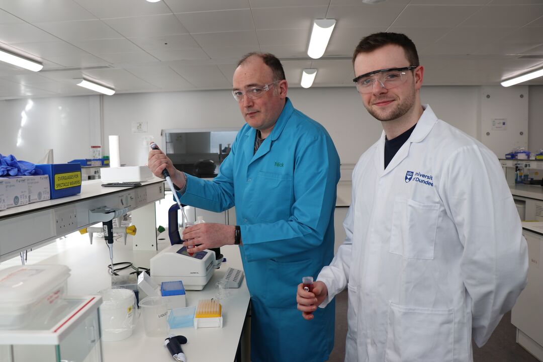 Two men standing at science lab bench, carrying out science experiments together. On the left, older man wears blue lab coat, on right younger man wears white lab coat.
