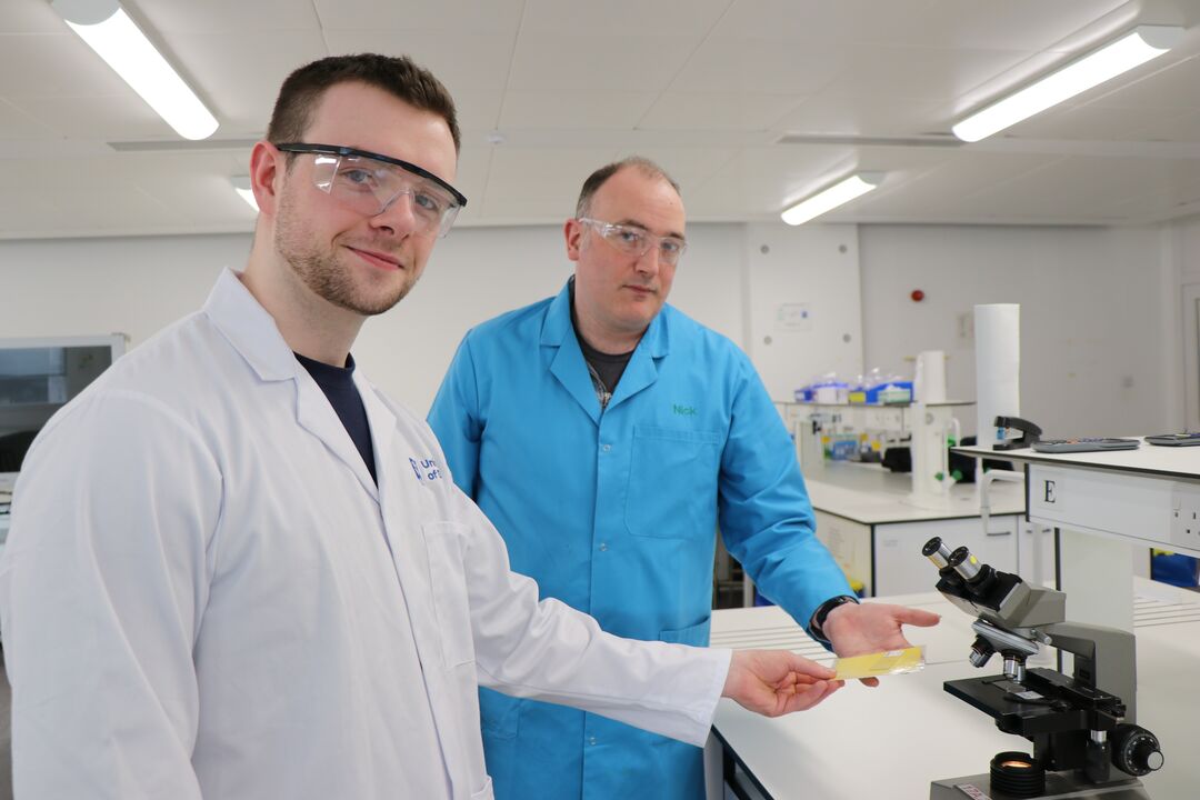 Two males in science lab wearing lab coats, standing at microscope, passing a sample disk between them, while looking at the camera