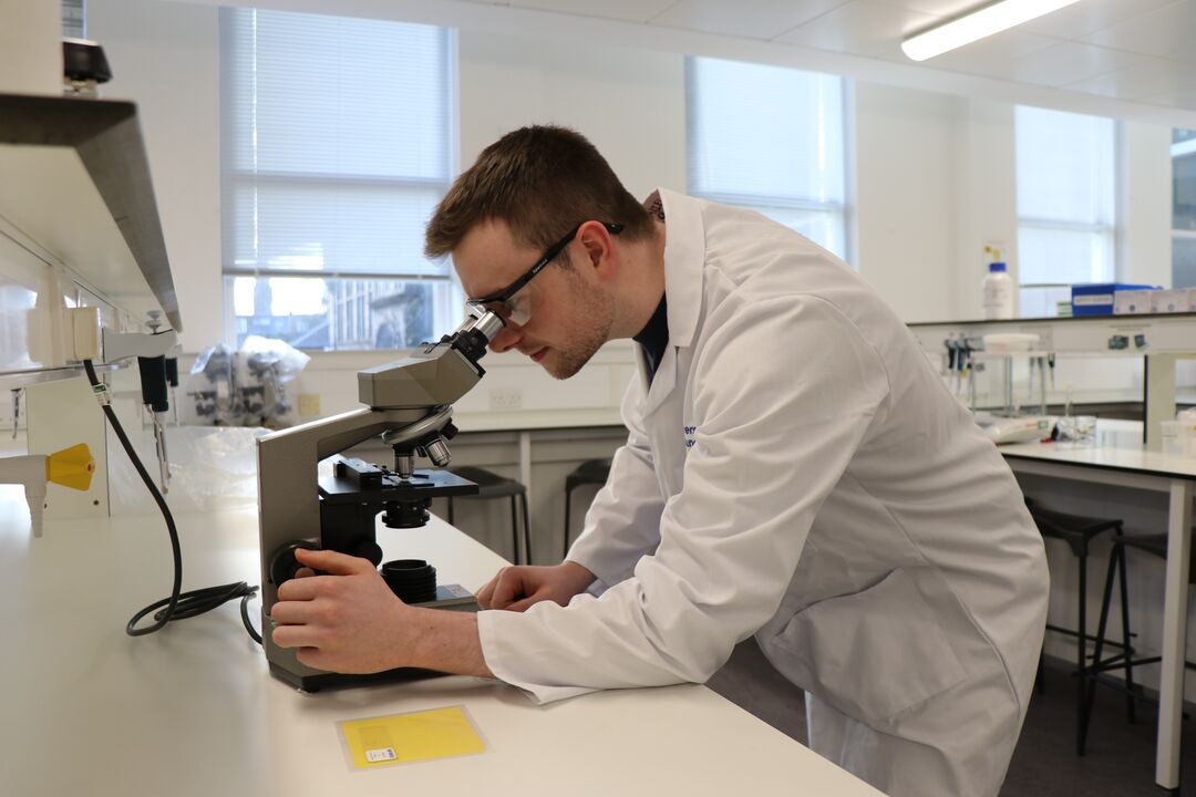Young man wearing white lab coat and protective eye wear, looking into microscope, in a science lab