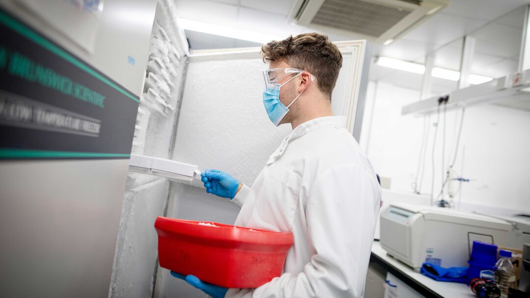 A person in a lab next to a large fridge holding a red plastic container