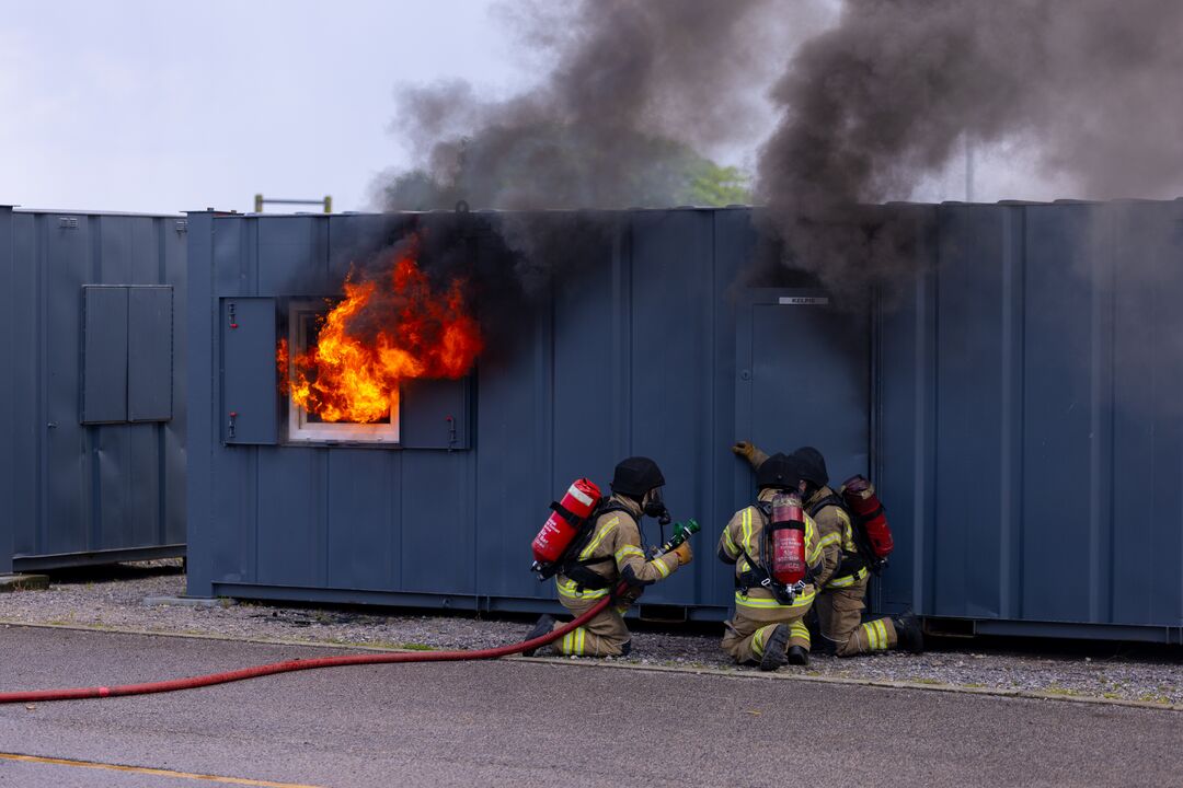 Shipping container with window on left which is broken and flames are coming out of it. Firefighters at door in centre holding hoses and spraying water in through the door.