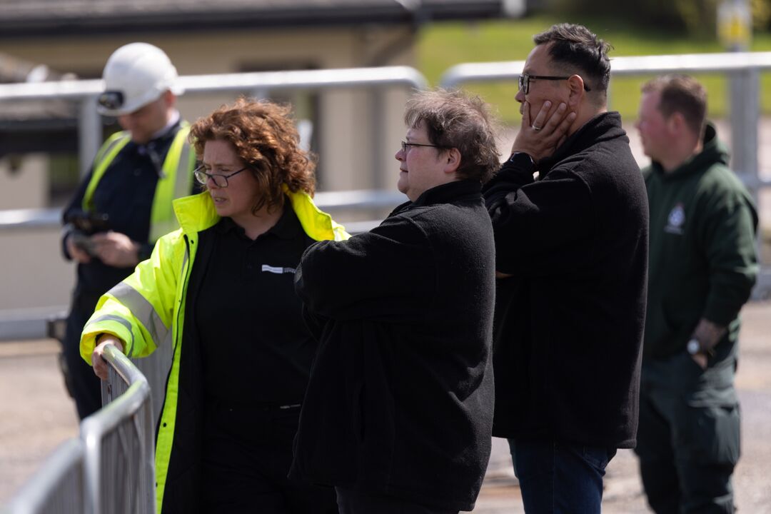 Three people standing at a fence looking over it at something out of shot. Two women, one wearing a high visibility jacket, and one man