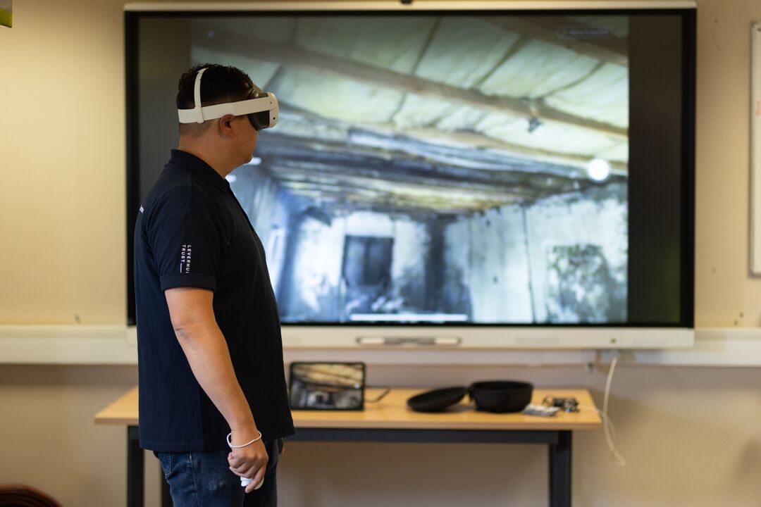 Man standing side on wearing a virtual reality headset over his eyes, with a large screen behind him displaying a room burnt out by fire