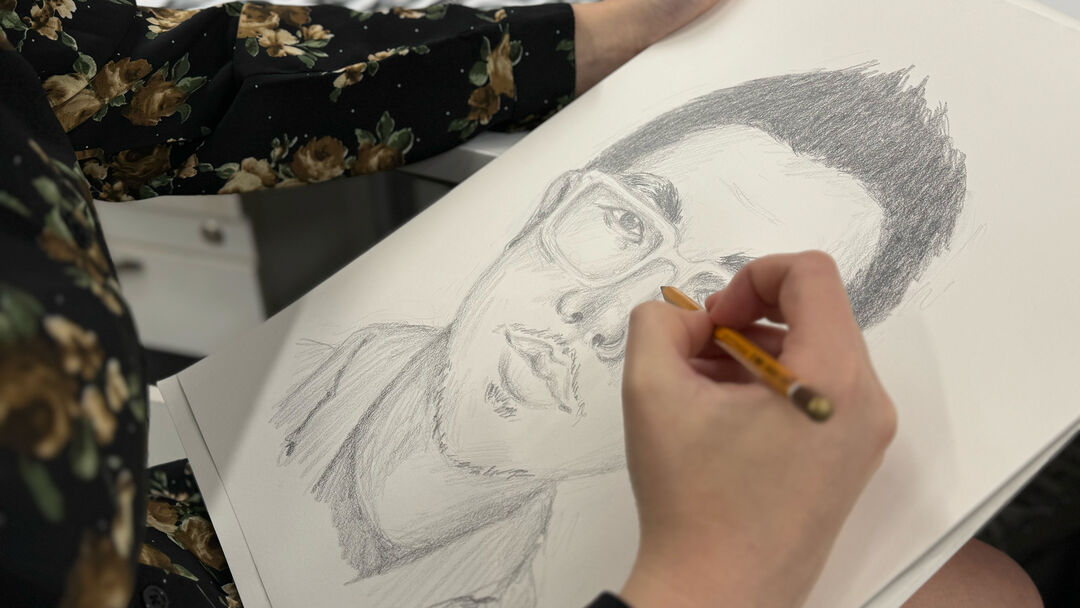 Close up of an unseen individual's hands sketching a face of a man with a pencil