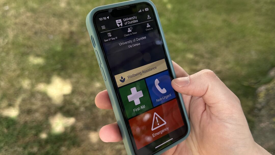 A closeup photo of an iPhone showing the SafeZone main interface. From top to bottom, there is a University logo with a menu icon to its left. On the next row are icons for "See it? Say it?", "Check in timer", and "Check in". There is then a large narrow button for "Well being assistance", then half width buttons for "First Aid" and "Non-Urgent". Finally at the bottom is a full width red button for "emergency"