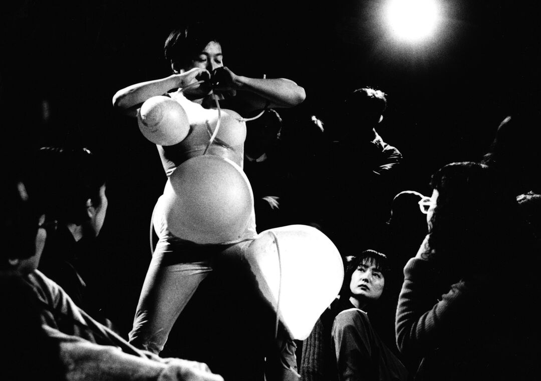 Tari Ito performance view black and white. Artist blowing inflatables around her body with tubes connected to her mouth creating enlarged spheres on her stomach, chest