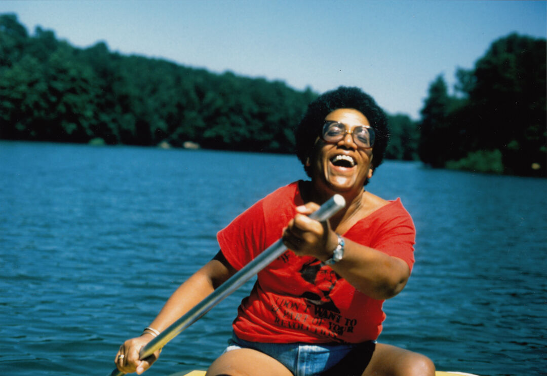Photo of Audre Lorde on a lake laughing. She is a black woman and is paddling, laughing, wearing afro hair, glasses and a red t-shirt