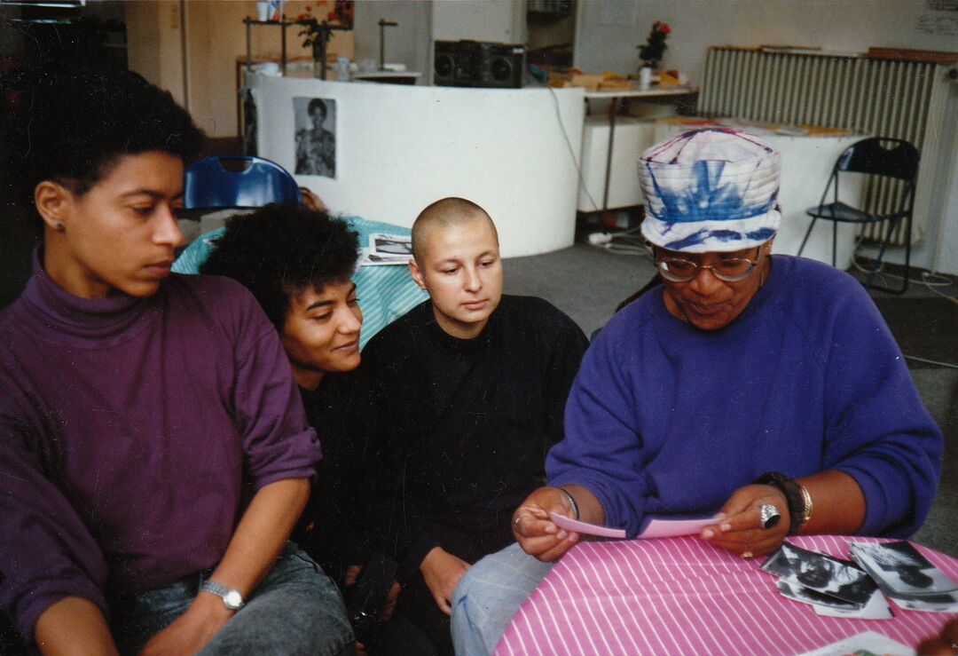 Four black people sit around a table as Audre Lorde looks through printed photographs. She wears a tie-dye hat and glasses. The others look closely at the photos.