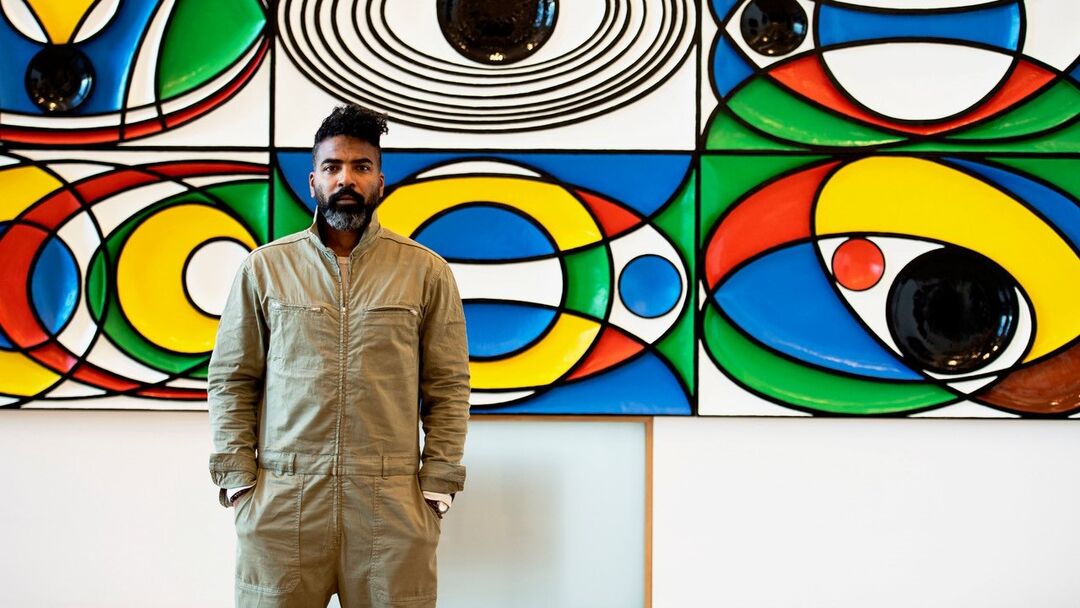 A man wearing khaki green overalls standing in front of a brightly coloured geometric patterned artwork