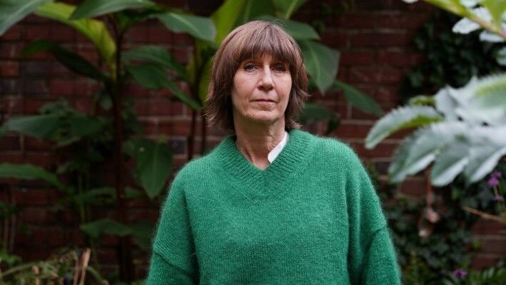 head and shoulders shot of woman wearing a green woolly v neck jumper, looking off into the distance, with large plants behind her