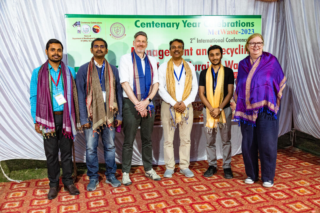 group of five males and one female, standing in a line in front of a large green sign with writing on it. The group are of mixed ethnicity and all are wearing Indian silk scarfs. 