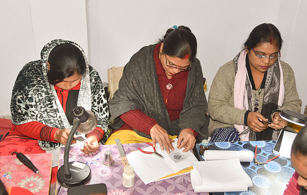 three women sat at a table with lots of craft materials and a light on it. The woman are all looking down towards their hands where they are holding crafting projects which they are working on