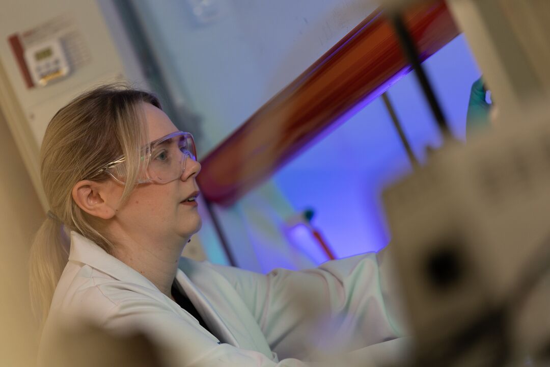 Woman in white lab coat and safety glasses, in a chemistry lab working with science equipment