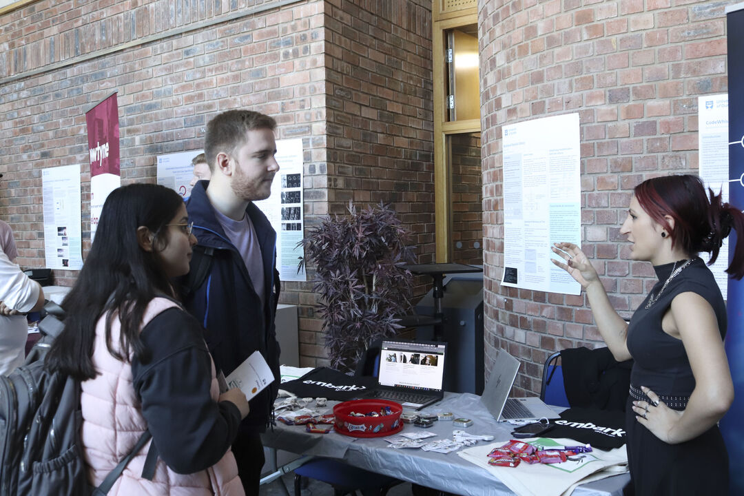 A male and a female stand at a desk while another female speaks to them, with one hand on her hip and the other in the air in front of her.