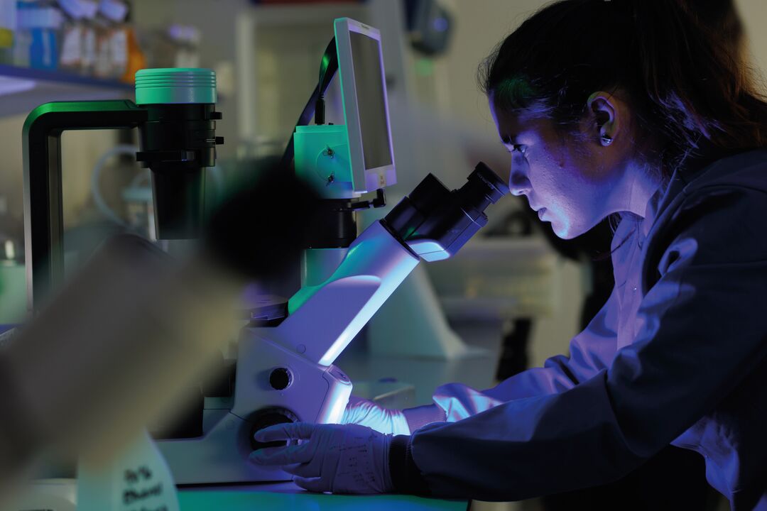 A woman looks into a microscope in a lab. The room is bathed in blue light.