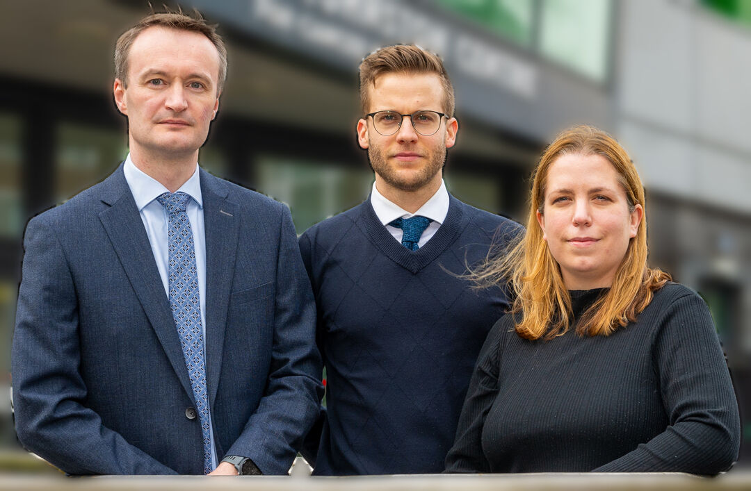 Group of three people wearing smart clothes looking and smiling at camera. Two males, centre and left, and a female on the right