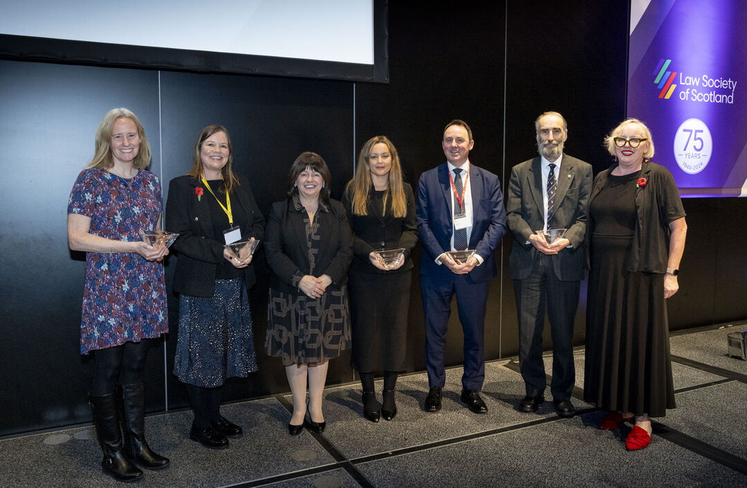 Group of seven adults standing in a line, wearing smart clothes, and five of them are holding glass awards