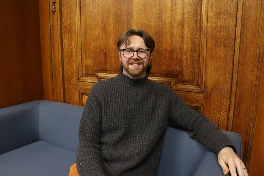 man with mid length dark hair sat on sofa, looking and smiling at camera