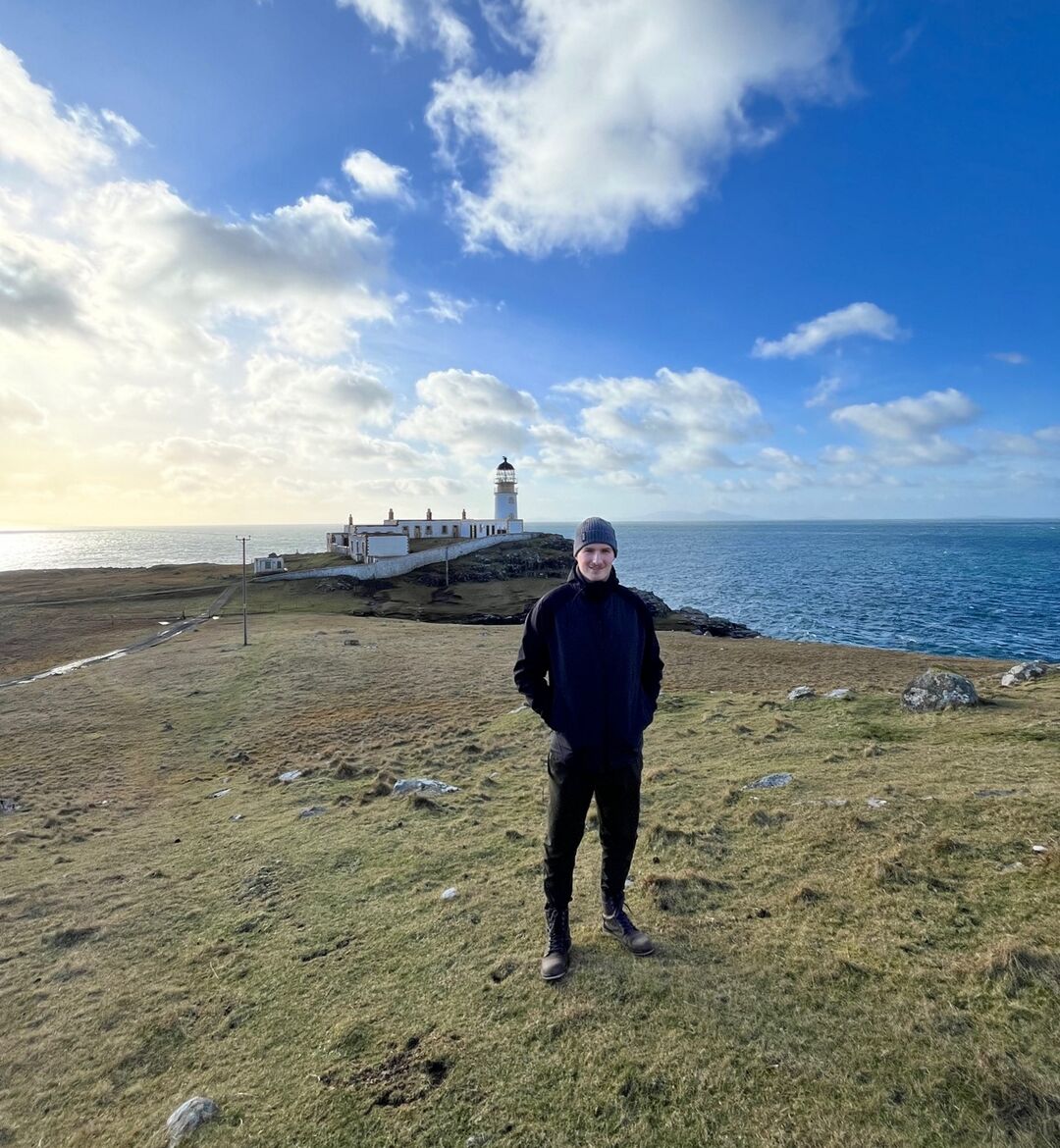 A man in dark clothing stands on a headland with a lighthouse behind him.
