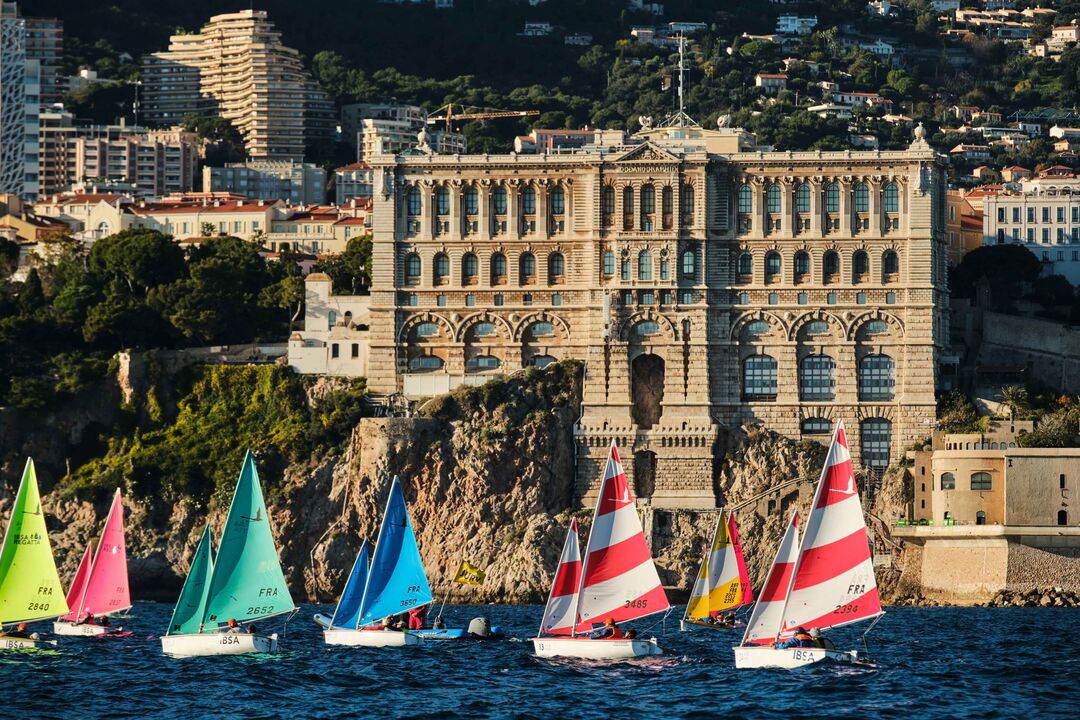 Several colourful sail boats taking part of the Navicap Challenge in Monaco with a stately building on shore in  the background
