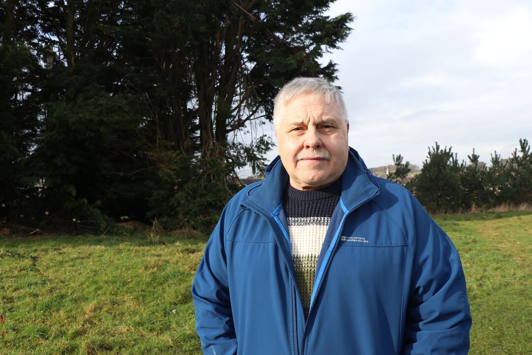 man with short grey hair, wearing an outdoors jacket, standing in a park with trees behind him, looking at the camera