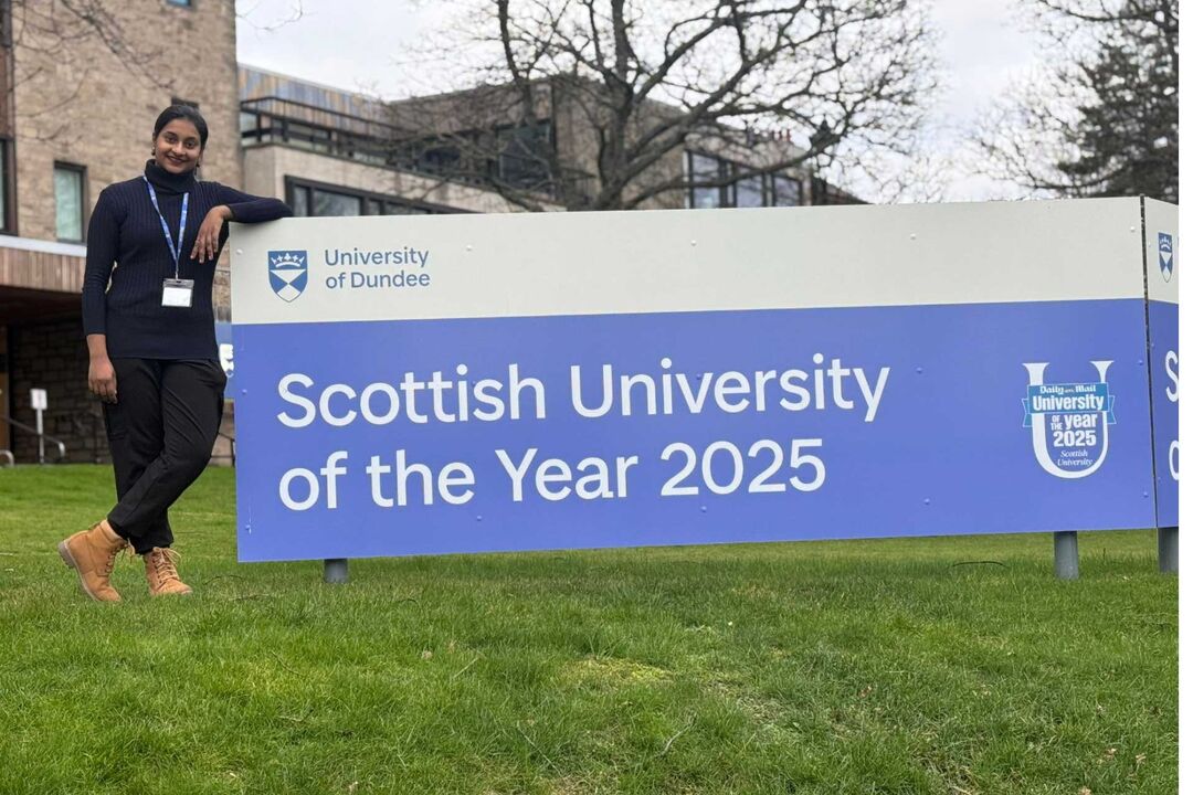 A woman dressed in black leans on a large blue sign that reads "Scottish university of the year 2025".