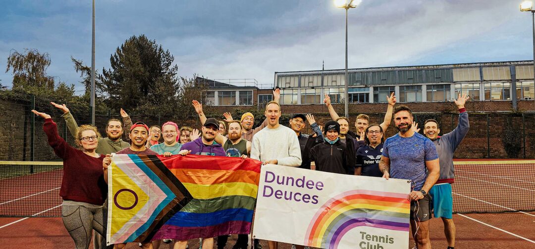 A group of people stand on an outdoor tennis court holding a large LGBTQ+ flag between them.