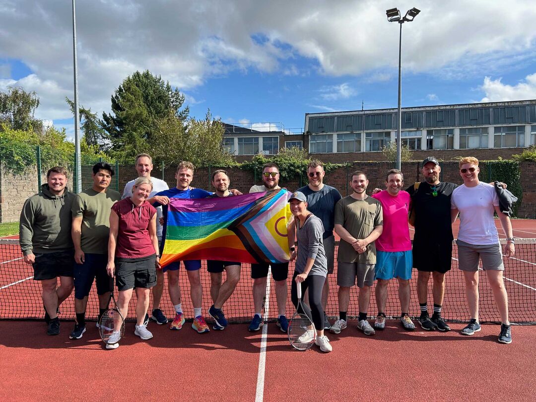 A group of people stand on an outdoor tennis court holding a large LGBTQ+ flag between them.