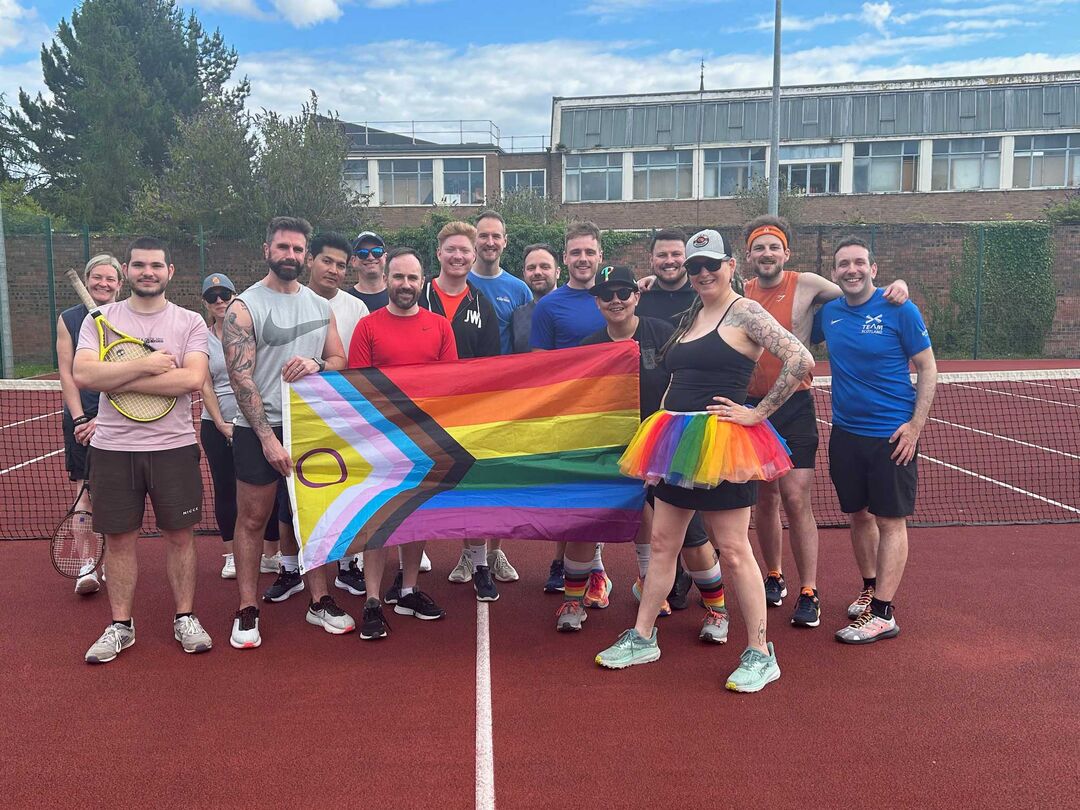 A group of people standing on an outdoor tennis court holding an LGBTQ+ flag between them
