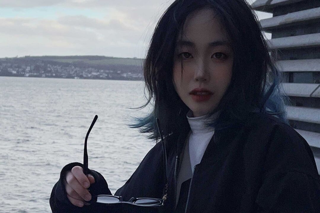 A girl with straight black hair holds a pair of sunglasses in her hand, there is water and a land mass behind her.