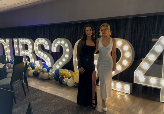 Two women in formal dresses stand in front of large lit up letters.