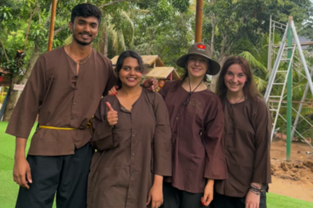 Four people stand together, all wearing brown shirts, there is jungle and a metal structure behind them.