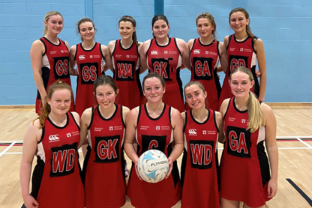 A team of sportswomen in read stand in two lines, one girl in the front holds a ball.