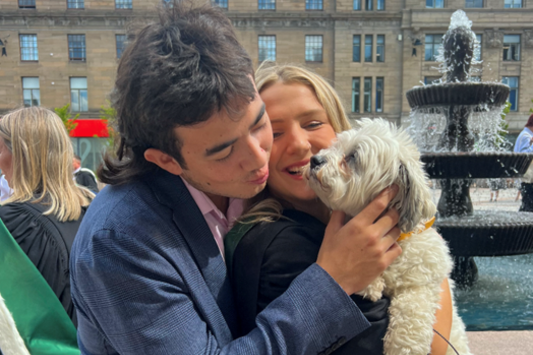 A man and woman stand together with a small white fluffy dog in their arms.