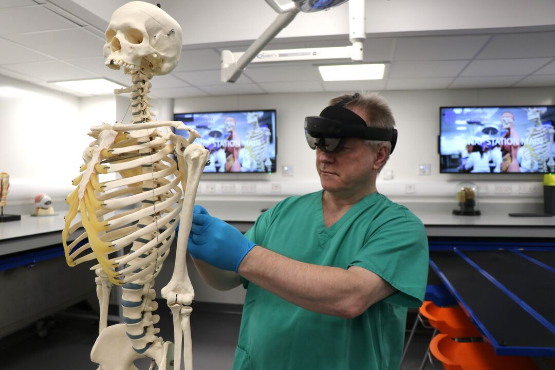 man wearing medical scrubs and a visor holds a needle up to the back of a skeleton, displaying a medical procedure