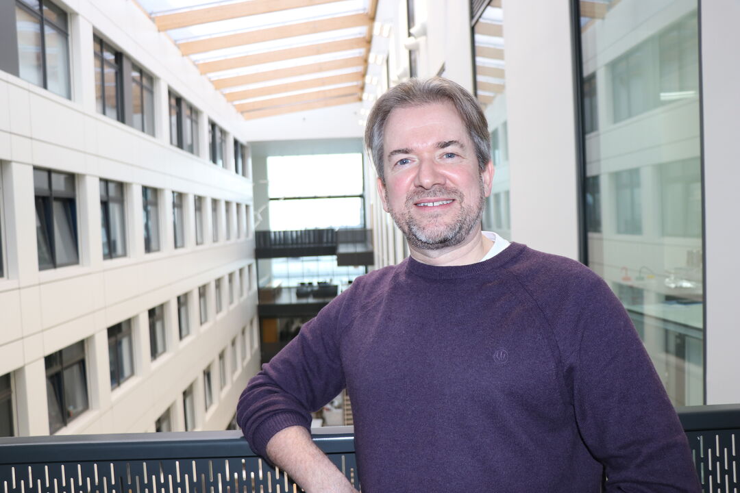 man wearing a purple jumper, standing on a balcony, with his arm resting on the edge. he is looking at camera and smiling, and a building can be seen behind him.